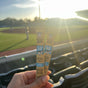 A hand holds two Brutus Bone Broth On The Go sticks at a stadium with empty seats, as sunlight shines over a baseball field and players in the background.
