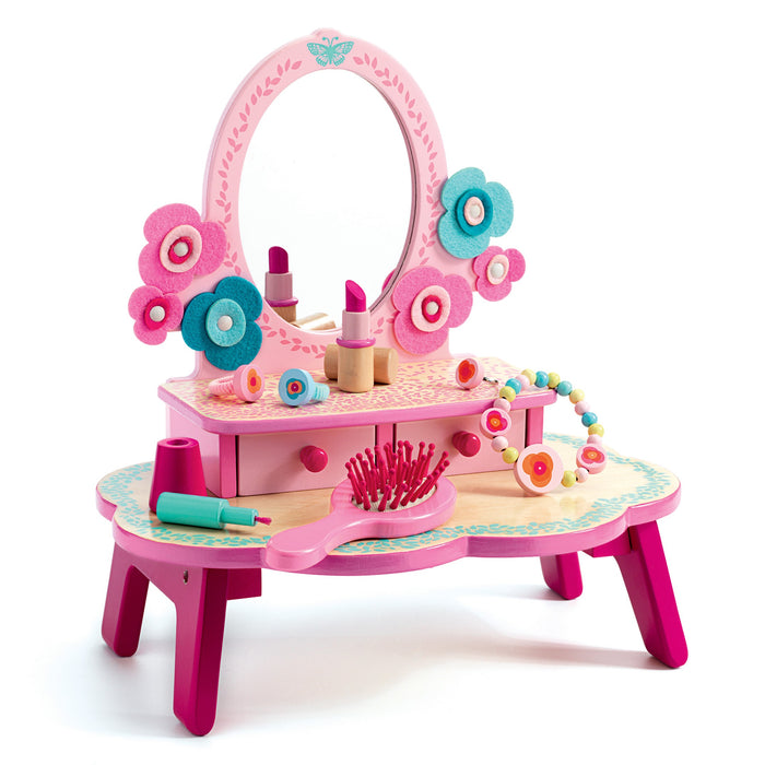 A colorful children’s vanity set with a pink-framed mirror, felt flower decorations, toy lipstick, nail polish, a beaded necklace, hairbrush, and a small drawer, all on a pink and teal wooden table.