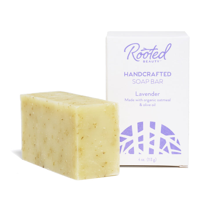 A rectangular beige bar of soap with flecks, placed next to a white box labeled Rooted Beauty Handcrafted Soap Bar, Lavender, Made with organic oatmeal & olive oil.