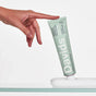 A hand reaches for a green tube of Davids Premium Toothpaste - Whitening + Antiplaque, which is standing upright on a white holder against a plain white background.
