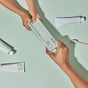 Two hands exchange a tube of Davids Hydroxi Whitening + Enamel Health Toothpaste with nano hydroxyapatite for Kids and Adults on a light green surface, surrounded by more fluoride-free toothpaste tubes and a toothbrush, emphasizing the brand’s dedication to natural whitening.