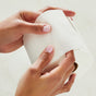 Two hands with light pink nails hold a Grove Co. 3-Ply Bamboo Toilet Paper roll, partially unrolled on a plain, light surface. The persons fingers pinch the paper gently, showcasing its texture and softness.