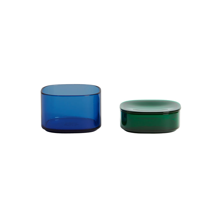 Two glass containers on a white background: one is a tall, cylindrical blue container, and the other is a shorter, rounded rectangular green container with a matching lid.
