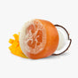 Explore Soapymanias Mango Coconut Loofah: a round, translucent handmade soap with an embedded loofah, mango slices, and half a coconut on a pristine white backdrop. Its orange hue and lighter bottom layer promise an eco-friendly cleanse.
