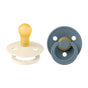 Two baby pacifiers are shown: one is cream-colored with a yellow nipple, and the other is blue-gray with a matching ring and yellow accent. Both have circular shields with ventilation holes.