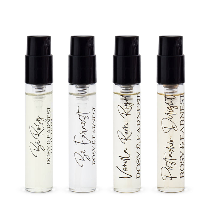 Four small glass perfume vials with black spray caps, each labeled with different scents: Be Rosy, Be Earnest, Vanilla Rum Rosy, and Patchouli Delight by Rosy & Earnest.