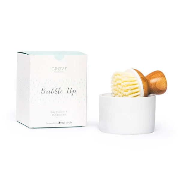 A wooden-handled dish brush with white bristles rests in a white ceramic dish next to a box labeled Grove Bubble Up Soap Dispenser & Dish Brush Set.