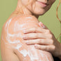 A woman with wavy hair smiles softly while applying Weleda Skin Food Body Wash to her bare shoulder, supporting her skin’s moisture barrier against a light green background.