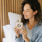 A woman with wavy hair and closed eyes smiles while holding a reed diffuser. She wears a striped shirt, denim jacket, and several rings, sitting on a bed with a wooden slat wall in the background.