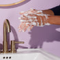A person uses Grove Co.s eco-friendly Hand Soap Sheets (40 Washes). Foamy white bubbles cover their hands over a white sink with brass fixtures, releasing a natural fragrance. The light purple wall in the background complements the serene setting.