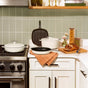 A modern kitchen with white cookware on the stove and counter, a sliced loaf of bread and knife on a wooden board, glass jars with pasta and grains, and a folded orange cloth on the counter.