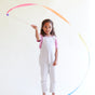 A young girl in light overalls and a pink shirt joyfully waves her Rainbow Streamer by Sarahs Silks against a white background, creating vibrant color arches in the air—an invitation to imaginative play.