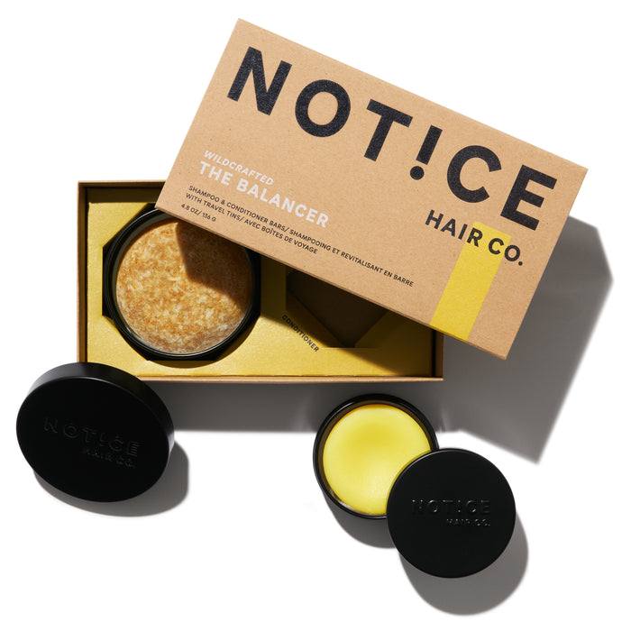 A brown cardboard box labeled NOTICE Hair Co. sits open, revealing a round solid shampoo bar inside. Two black containers, one open showing a yellow balm, are beside the box on a white surface.