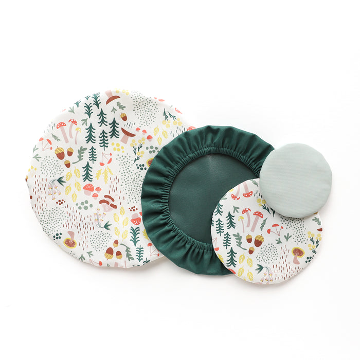 Four reusable fabric bowl covers in various sizes are arranged on a white background. Two covers feature a woodland pattern, while the others are solid dark green and light blue-green.