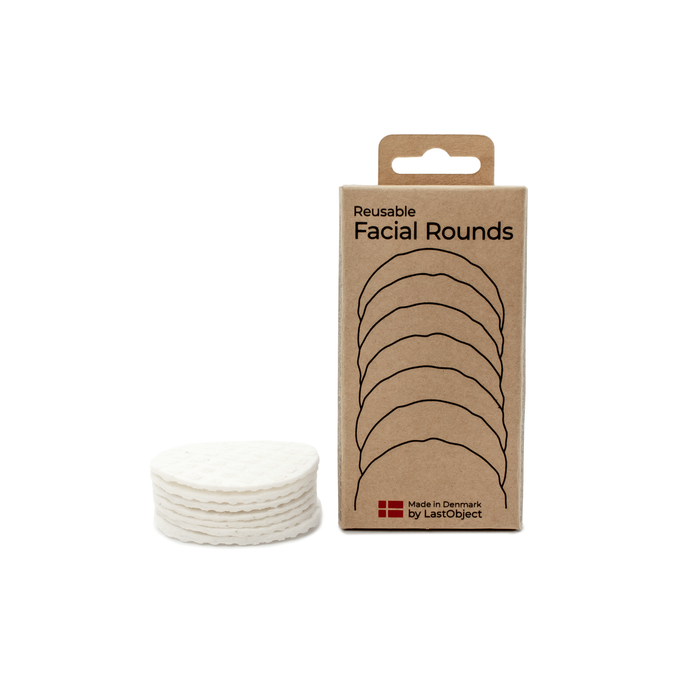 A stack of white reusable facial rounds is placed next to a brown cardboard box labeled Reusable Facial Rounds. The box has an illustration of stacked rounds and text indicating its made in Denmark by LastObject.