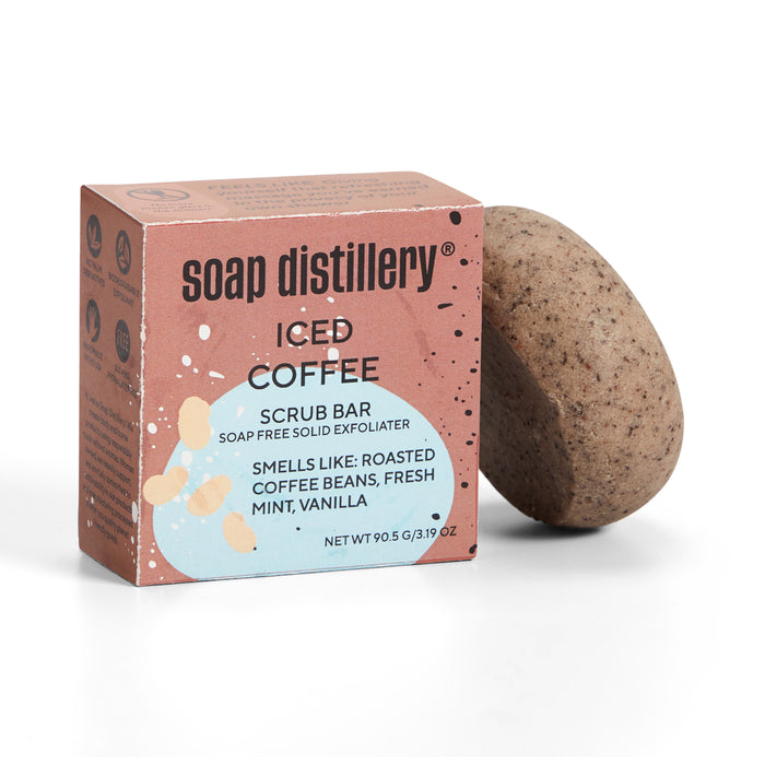 A Soap Distillery Iced Coffee Scrub Bar next to its box. The box describes the scent as roasted coffee beans, fresh mint, and vanilla. The scrub bar is speckled and oval-shaped.