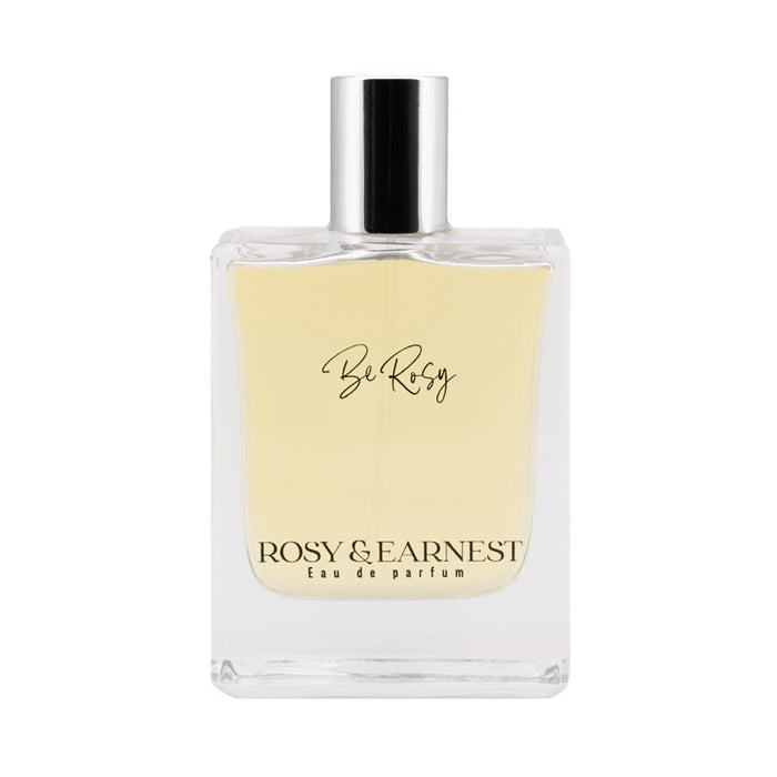 A clear glass bottle of perfume labeled Be Rosy with Rosy & Earnest Eau de Parfum printed at the bottom, filled with light yellow liquid and topped with a silver cap.