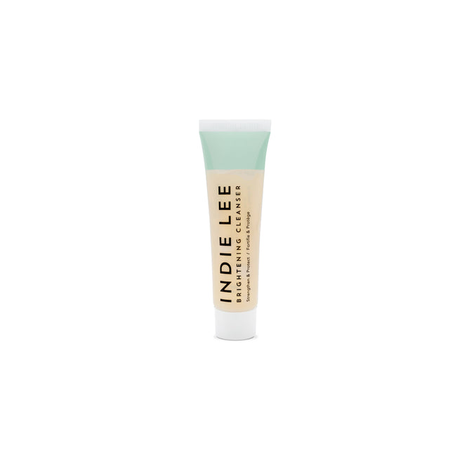A small tube of Indie Lee Brightening Cleanser with a pale green cap and clear body, showing the light-colored liquid inside. The product name is printed vertically in black letters.