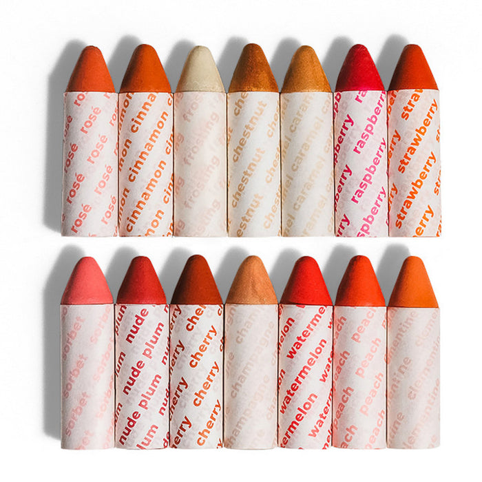 Twelve crayon-shaped lip balms with pointed tips, each wrapped in white paper printed with the shade name, are arranged in two rows by color, ranging from reds and pinks to nudes and browns.