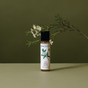 A small bottle of Koala Eco natural dish soap stands on a green surface, with a sprig of white flowers and green leaves placed behind it against an olive-green background.