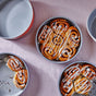 Round pans filled with cinnamon rolls, some drizzled with white icing, sit on a light pink tablecloth. One pan is nearly empty, while others hold neatly arranged rolls.