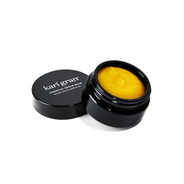 A small, round black jar with the lid off, revealing a yellow balm inside. The lid, placed beside the jar, reads kari gran essential balm in white text.