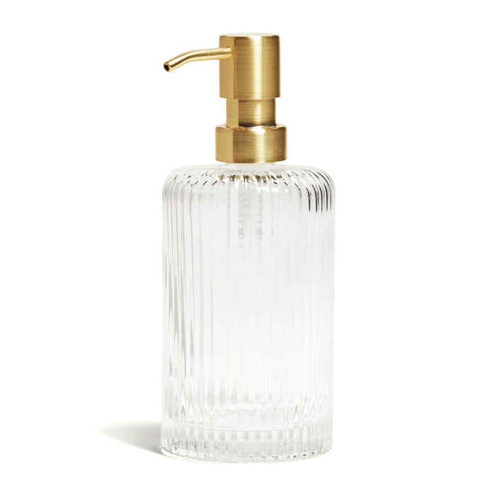 The Grove Co. Eves of Enchantment is a limited edition clear ribbed glass hand soap dispenser with a gold pump nozzle and lid, perfect for festive scents. This eco-friendly dispenser adds elegance to any bathroom decor and stands out against its plain white background.