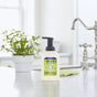 A bottle of Mrs. Meyer’s Clean Day foaming hand soap sits on a white kitchen countertop next to a small potted plant, with a sink and green cloth in the background.