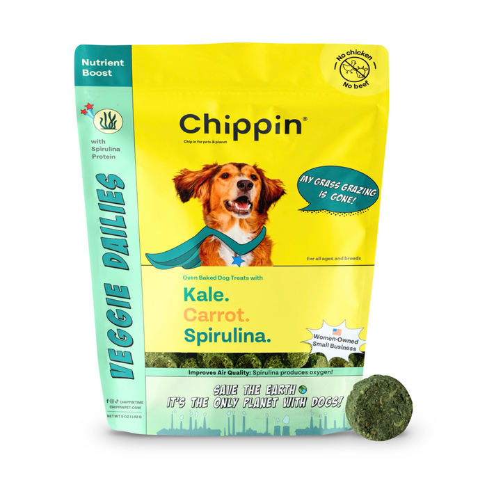 A bag of Chippin Veggie Dailies dog treats with a photo of a happy dog wearing a cape. The packaging promotes kale, carrot, and spirulina ingredients and highlights no chicken/beef, nutrient boost, and eco-friendly benefits.