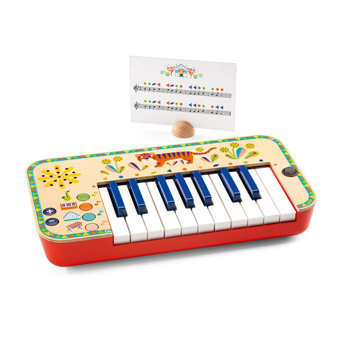 A colorful toy piano with eight keys, decorative patterns, and buttons on the left side. A music sheet with notes and a small wooden mallet are positioned behind the piano.