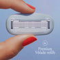 A hand with red-painted nails holds a razor blade refill against a light blue background. The refill has five blades, and text reading 