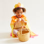 A child wearing a yellow and orange costume and a paper crown sits on the white floor, holding a wooden egg from Sarahs Silks. A wicker basket full of Montessori toys is in front of them.