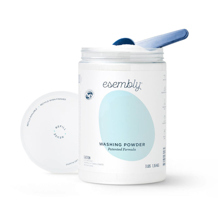 A container of Esembly washing powder with the lid off, showing a blue scoop resting inside the white powder. The label reads “Esembly Washing Powder Patented Formula.”.