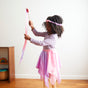 A young girl with curly hair, dressed in purple and pink, twirls Sarahs Silks Heart Streamer ribbon wand energetically. Standing on a wooden floor near a small toy shelf against a plain white wall, she hones her gross motor skills with every graceful move.