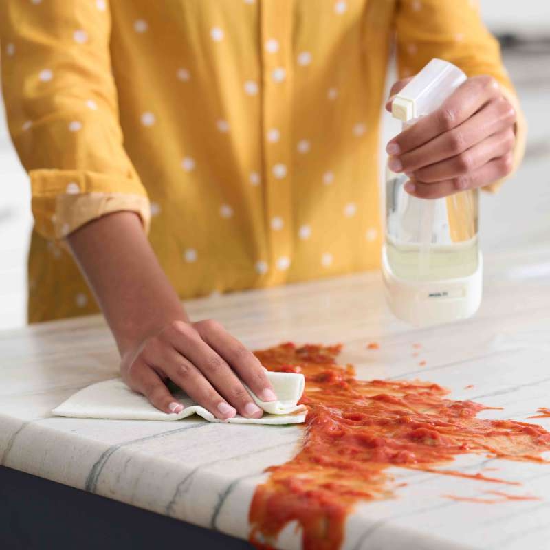 Wearing a yellow shirt, someone cleans spilled sauce on a marble countertop using Grove Co.s Multi-Purpose Cleaner Concentrate. The bio-based ingredients and recyclable packaging make the clean-up feel both eco-friendly and efficient.