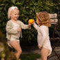 Two young children wearing diapers and light-colored clothing stand outdoors by a wooden chair. One child hands an orange to the other, and greenery fills the background.