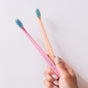 A hand with light-colored nail polish holds two toothbrushes, one pink and one orange, each with blue and white bristles, against a plain white background.