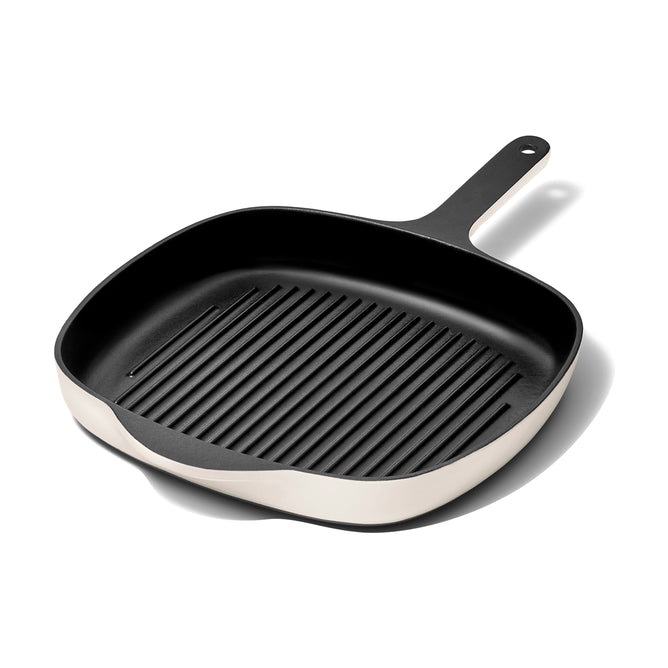 A square, black cast iron grill pan with raised ridges, a light-colored exterior, and a long handle with a hole at the end, shown on a white background.