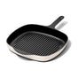 A square, black cast iron grill pan with raised ridges, a light-colored exterior, and a long handle with a hole at the end, shown on a white background.