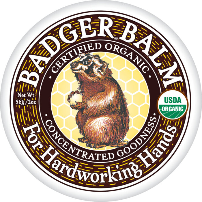 A round tin lid labeled Badger Balm For Hardworking Hands features an illustration of a badger holding a white object, with honeycomb background, and organic certification seals.