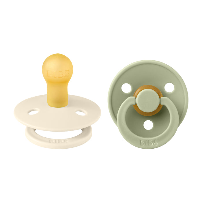 Two baby pacifiers are shown, one in cream and the other in sage green. Both have round rubber nipples and ring handles, with the brand name BIBS embossed on them.