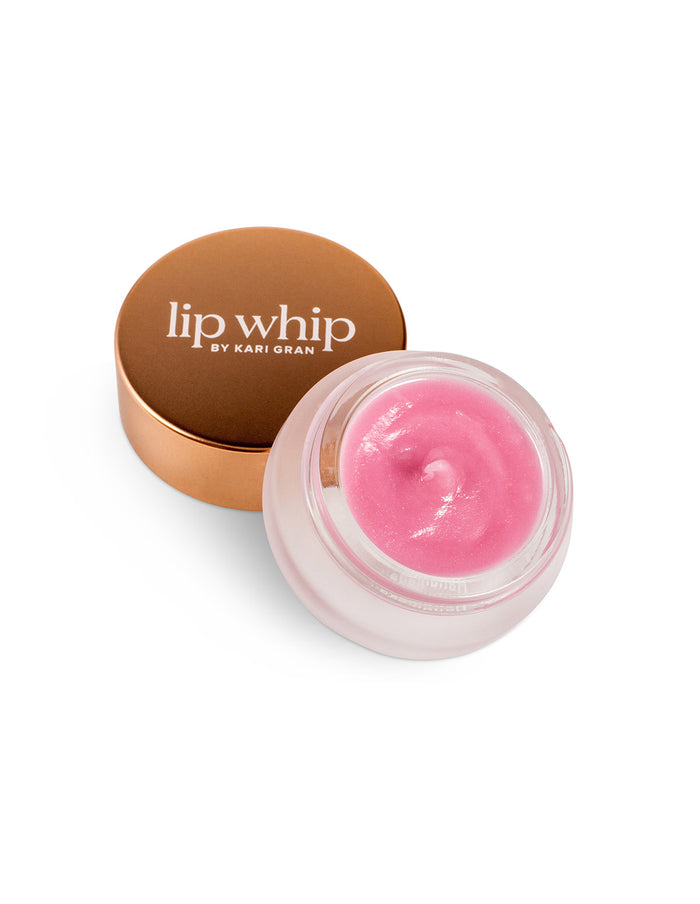 An open jar of Kari Gran Lip Whip shows a smooth, pink lip balm inside. The gold lid, placed beside the jar, displays the white text “lip whip BY KARI GRAN” on top. The background is white.