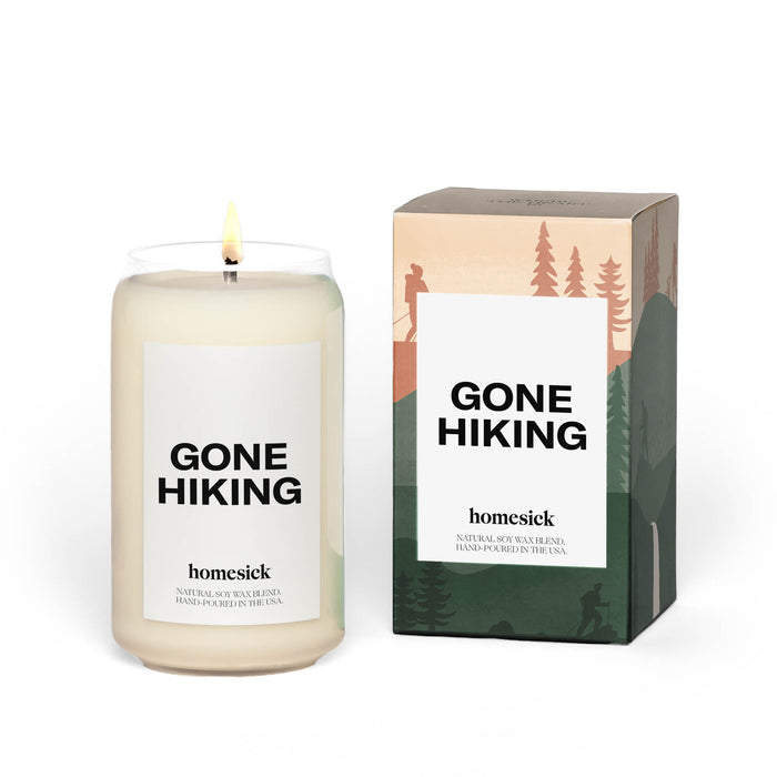 A lit candle in a clear glass labeled GONE HIKING sits next to its box, which features silhouettes of hikers and trees. The candle is branded homesick and described as natural soy wax and hand-poured in the USA.