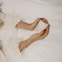 A person on a white bed, in a feather-trimmed garment, holds a PAUME Overnight Hand and Foot Hydration Mask jar. Their red nails and gold bracelets glisten as they prepare for hydration with the nourishing power of shea butter.
