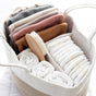 A woven basket filled with folded baby clothes, rolled white towels, a wooden baby brush, and neatly stacked disposable diapers with yellow lines.
