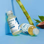 Two small bottles of Grove Co. Glass Cleaner Concentrate are placed on a blue surface, one upright and one lying down. A bamboo stalk and a halved fig are in the background.