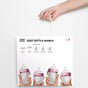 A hand holds a white shopping bag labeled comotomo BABY BOTTLE BUNDLE with images of four pink-topped baby bottles and a list of bundle contents printed on the front.