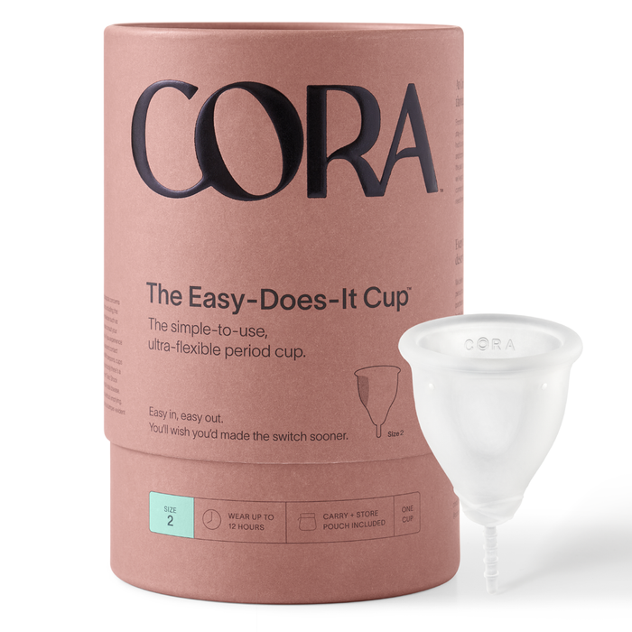 A cylindrical container labeled CORA The Easy-Does-It Cup stands next to a clear menstrual cup. The packaging is pink with black text and includes details about the product and its size (Size 2).