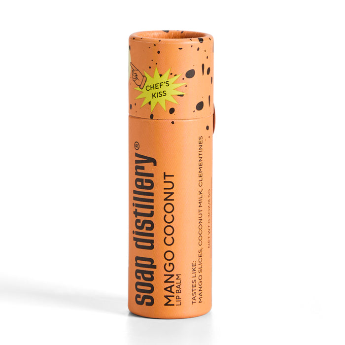 An orange tube of Soap Distillery Mango Coconut lip balm stands upright. The label features black text and a green burst with the words Chefs Kiss. Flavor notes include mango slices, coconut milk, and clementines.