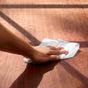 A hand uses Bioms All-Purpose Wipe Refills, made from natural ingredients, to clean a polished warm brown wooden surface while sunlight casts gentle diagonal shadows.